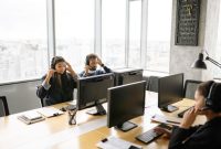 Seorang agen pusat layanan alih daya sedang mendengarkan keluhan pelanggan dengan cermat sembari mencatat data pada sistem komputer kantor.