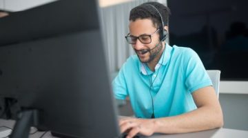 Seorang agen alih daya layanan pelanggan sedang melayani pertanyaan konsumen dengan ramah dan profesional menggunakan headphone di kantor pusat operasional.